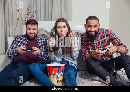 Three young friends playing computer games and drinking beer on the floor in living room Stock Photo