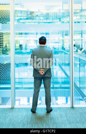 Businessman standing at atrium window with hands behind back Stock Photo