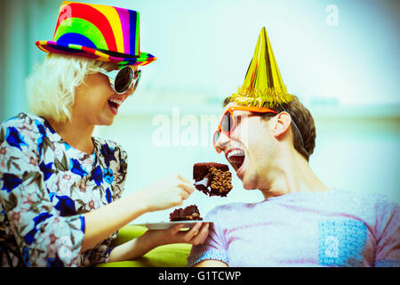 Playful couple in party hats and sunglasses eating chocolate cake Stock Photo