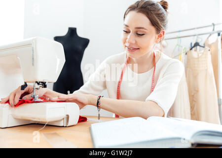 Smiling beautiful young woman seamstress sews on the sewing machine Stock Photo