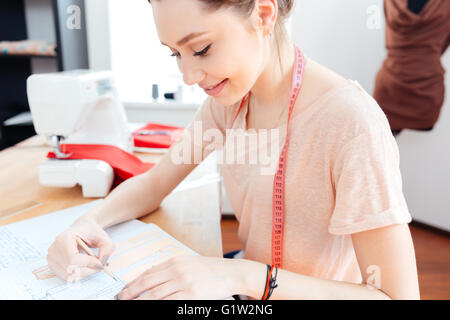 Happy attractive young woman seamstress sitting and drawing patterns in notebook Stock Photo