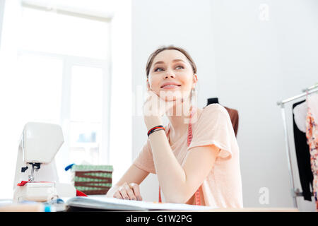 Smiling attractive young woman seamstress working with sewing machine and thinking Stock Photo