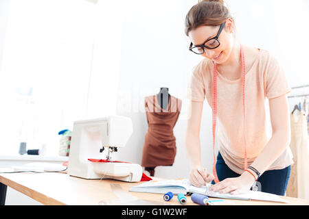 Happy cute young woman seamstress in glasses standing and drawing in workshop Stock Photo