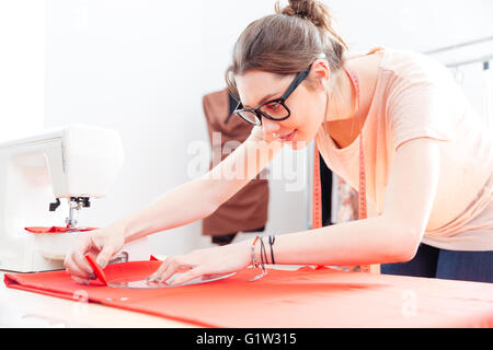 Focused attractive young woman tailor standing and drawing on red textile material Stock Photo