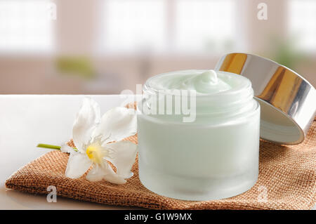 Glass open jar with facial or body cream on burlap. With lid and flower.Windows background. Front view. Stock Photo