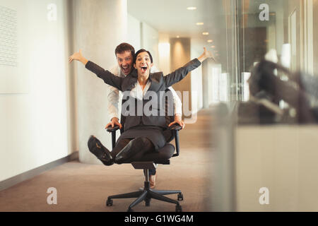 Playful businessman pushing exuberant businesswoman down corridor in office chair Stock Photo