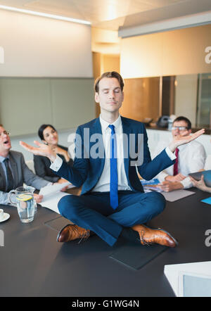 Colleagues watching zen-like businessman meditating in lotus position on conference table Stock Photo