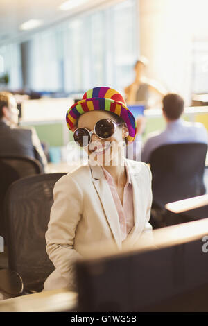 Portrait of smiling businesswoman in silly sunglasses and striped hat working in office Stock Photo