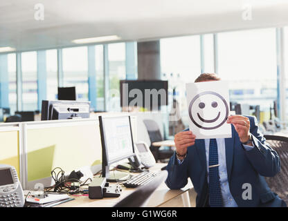 Portrait of businessman holding smiley face printout over his face in office Stock Photo