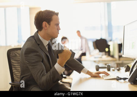 Businessman doing biceps curls with dumbbell at computer in office Stock Photo
