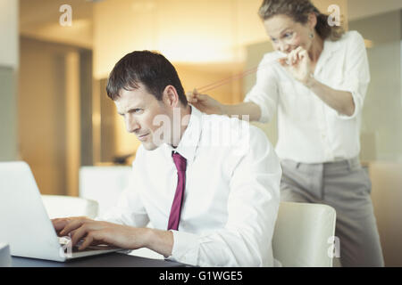 Mischievous businesswoman aiming rubber band at unsuspecting businessman working at laptop Stock Photo