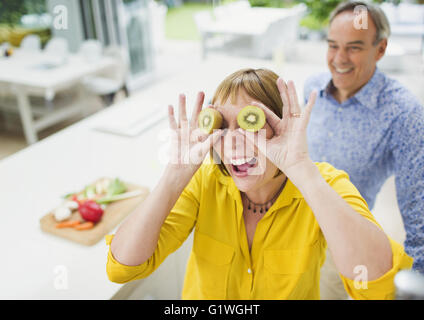 Portrait playful mature woman covering eyes with kiwi slices Stock Photo