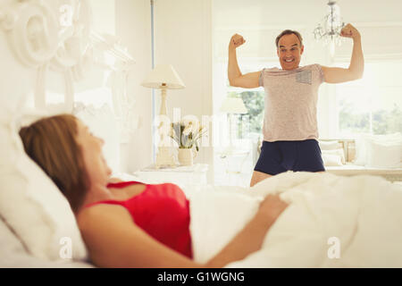 Playful mature man flexing muscles for wife in bedroom Stock Photo