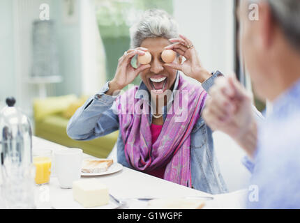 Playful mature woman covering eyes with eggs at breakfast table Stock Photo