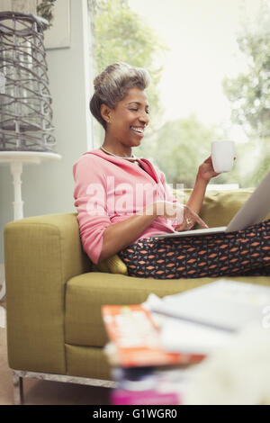 Smiling mature woman using laptop and drinking coffee on sofa Stock Photo