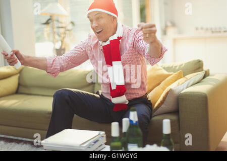 Enthusiastic mature man in hat and scarf watching TV sports event on living room sofa Stock Photo