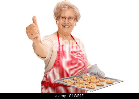 Mature lady holding a tray with chocolate chip cookies and giving a thumb up isolated on white background Stock Photo