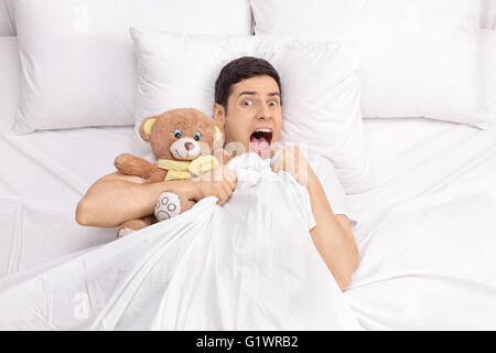 Top view of a terrified young man laying in bed with a teddy bear and screaming isolated on white background Stock Photo