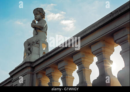 Little boy angel sculpture sits on the bridge construction and gesticulates. Blue sky background Stock Photo