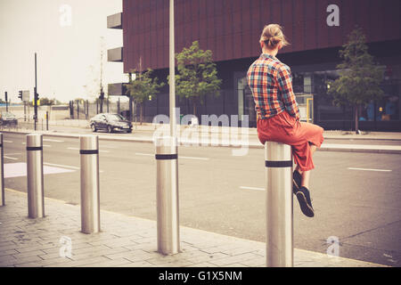 A young woman is sitting on a bollard by the roadside in the city Stock Photo