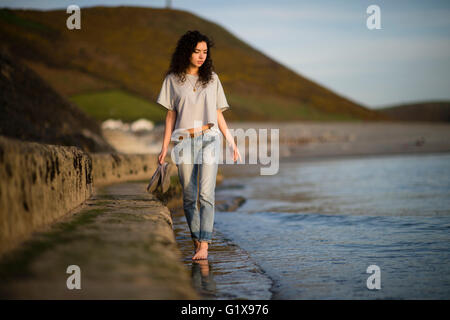A young attractive slim brunette woman girl  walking outdoors by the sea holding her espadrilles outside on a warm evening. UK Stock Photo