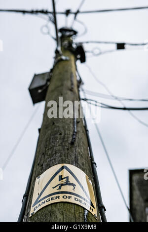 Old wooden telegraph pole seen next to a cottage showing a standard warning sign and both power and communication cables installed. Stock Photo