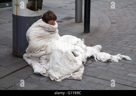 Homeless & homelessness & rough sleeper; living on the streets, doorway, sleeping, poverty, unemployment, welfare, beggar, tramp. A man seated, wrapped in a worn and torn duvet, on the pavement in poor condition in Liverpool One, Merseyside, UK Stock Photo