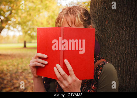 A young woman is in the park on a sunny autumn day and is hiding behind a book Stock Photo