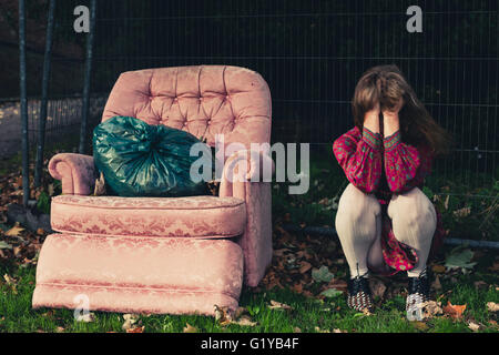 A sad young woman is sitting by an armchair outside on the grass Stock Photo