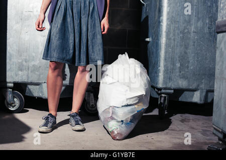 A young woman is standing next to some large bins with a bag of rubbish Stock Photo