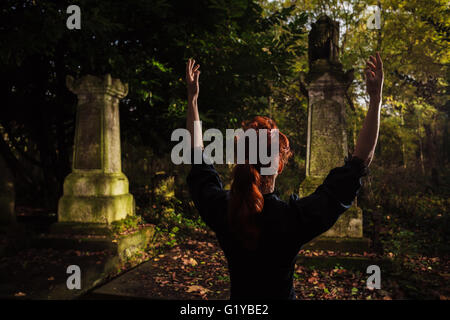 A redhead woman or witch is raising her arms and performing a ritual by a grave Stock Photo