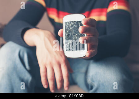 A young man is sitting on a sofa and is holding a broken smart phone with a cracked screen Stock Photo