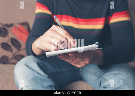 A young man is sitting on a sofa and is writing on the back of an envelope Stock Photo