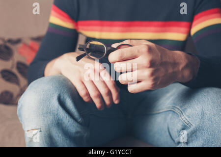 A young man is sitting on a sofa and is holding a pair of glasses in his hand Stock Photo