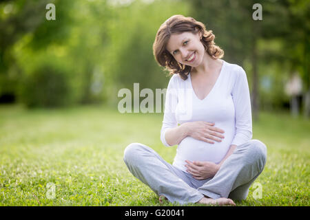 Portrait of happy young pregnant model sitting with crossed legs on grass lawn and looking at camera. Smiling future mom Stock Photo