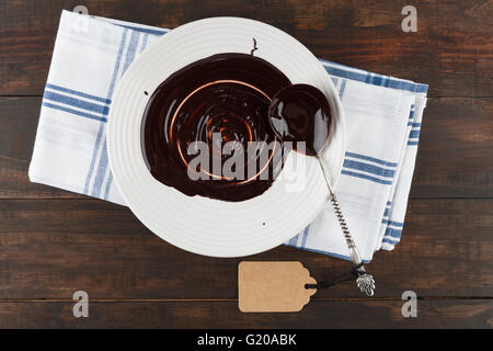 Vintage sliver spoon covered in melted chocolate on white plate and wooden table with gift tag. Top view. Stock Photo