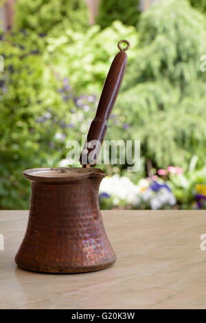 Old brown coffee pot with wooden handle on a table over green garden background with flowers Stock Photo