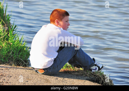 A boy, deep in thought, sits on a bank of the DesPlaines River as he gazes along the river's surface. Joliet, Illinois, USA. Stock Photo