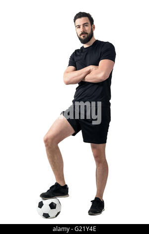 Football player standing on the ball with one leg posing with crossed arms. Full body length portrait over white background. Stock Photo