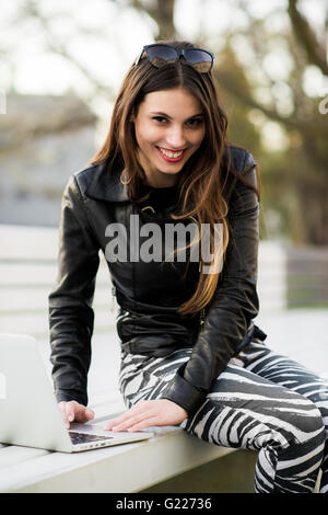 Girl works with laptop computer sitting on a bench in the park looking to camera and smiling Stock Photo