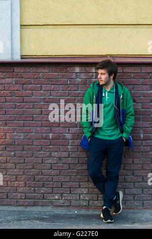 Common young man wearing green t-shirt jacket and jeans against a red brick wall looking sideways Stock Photo
