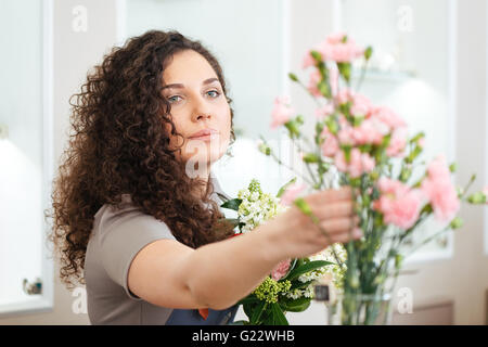 Beautiful concentrated young woman florist taking flowers and making bouquet in shop Stock Photo