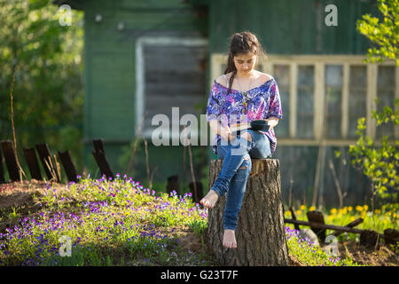 Teen girl reads book sitting on a stump in the yard in the village. Stock Photo