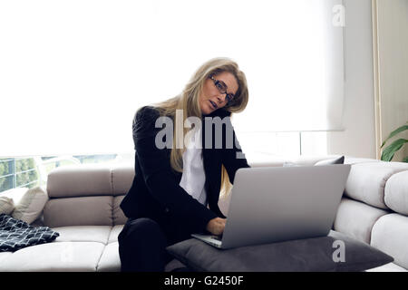 Middle-aged and attractive business woman making a phone call while working on laptop computer. Modern home office in bright int Stock Photo