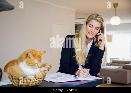 A middle aged woman wearing a suit happily talks on the phone while her pet cat sits atop the counter beside her in a wicker basket. Stock Photo
