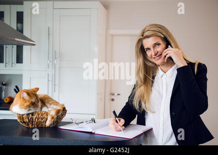 Attractive middle-aged business woman using a phone and taking notes. A ginger colored cat lying in a wicker basket next to her on the counter. Stock Photo