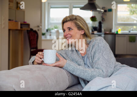 Blond woman relaxing at home on her couch with a hot beverage. Medium shot. Stock Photo