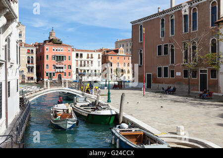 Tourists relaxing on Campo San Vio on the Grand Canal, Venice, Italy while a luggage transport boat passes by on a side canal Stock Photo