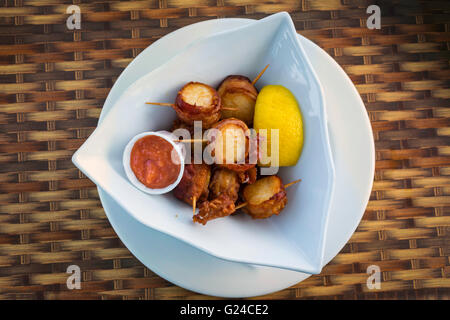 Bacon wrapped scallops with a slice of lemon and some seafood sauce in a serving dish. Stock Photo