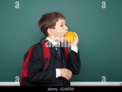 school boy eat orange in black suit on green chalkboard background with red backpack, education concept Stock Photo